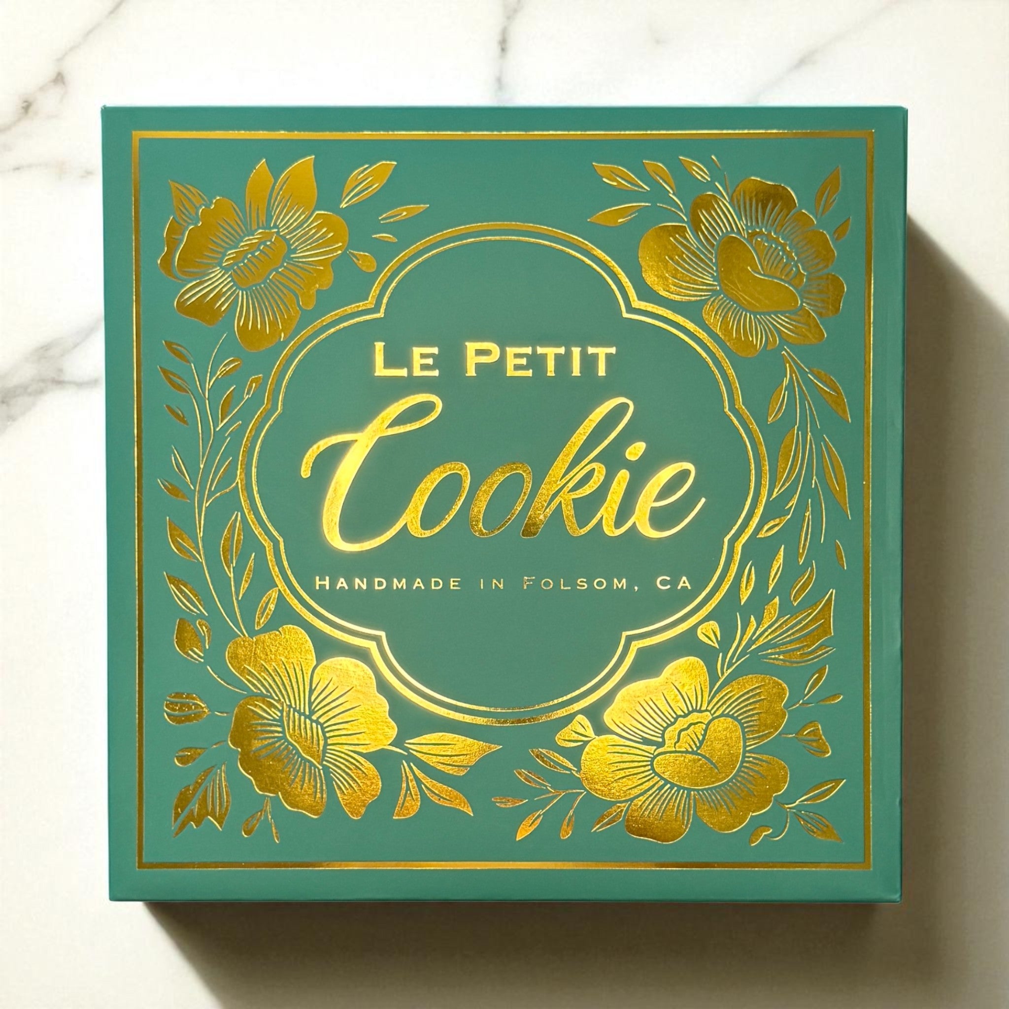 The exterior of the regular assortment cookie box from Le Petit Cookie featuring a teal box with gold writing that reads "Le Petit Cookie Handmade in Folsom, CA"