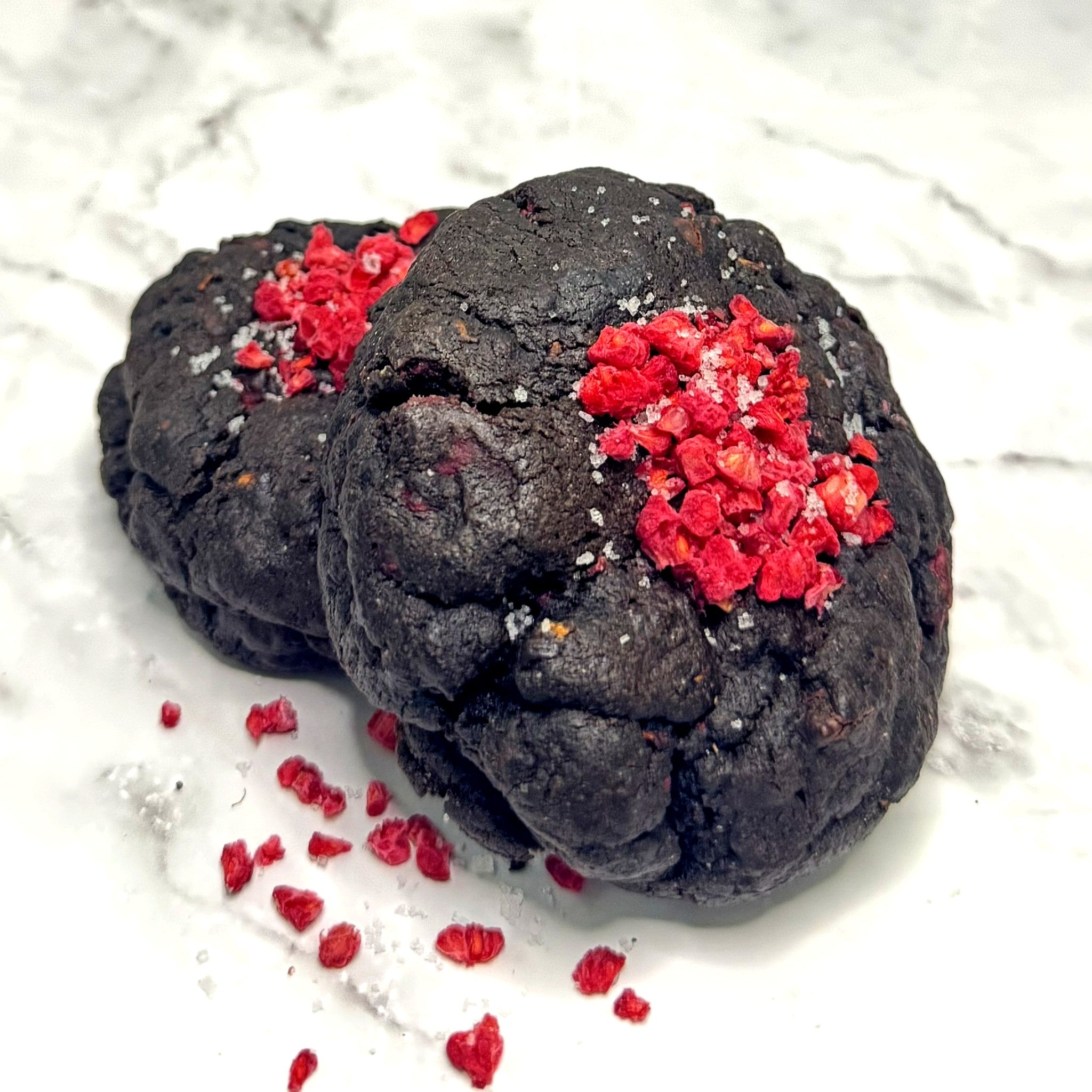 Two rich dark chocolate Midnight Berry Cookies from Le Petit Cookie stacked on top of each other on a marble countertop.